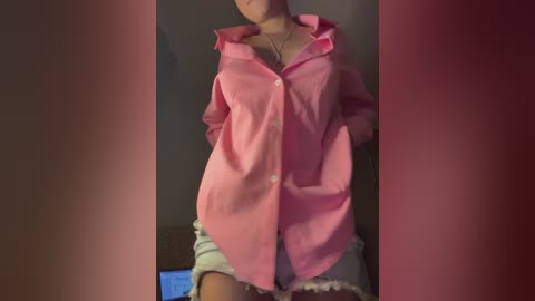 A video of a woman with light skin, wearing a pink button-up shirt, white shorts, and a necklace, standing indoors against a dark background.