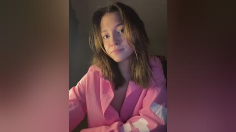 Video of a young woman with wavy, shoulder-length brown hair, wearing a pink robe, sitting indoors against a blurred background, with a neutral expression.