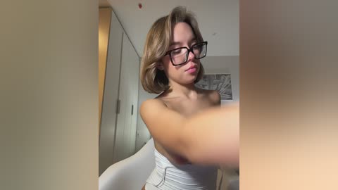 Media: Video of a young, light-skinned woman with shoulder-length, blonde hair and black-rimmed glasses, wearing a white top, standing in a modern, minimalist room with beige walls and a white chair.