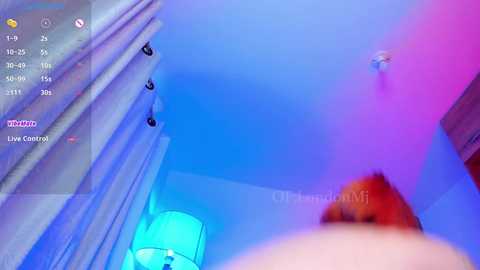 A video of a dimly lit, modern bedroom with a purple and blue light theme. The image is slightly out of focus, showing a person's face and upper body in the background.