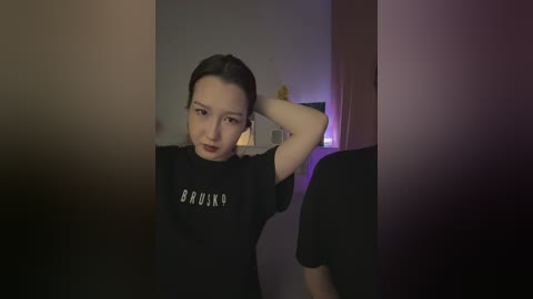 Media: A video of a young East Asian woman with fair skin, dark hair pulled back, wearing a black t-shirt with \"BROSKA\" in white letters. She stands in a dimly lit room with blurred objects in the background.