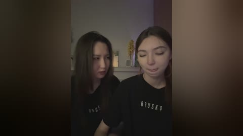 Media: Video of two young East Asian women with long black hair, wearing matching black t-shirts, standing close together, one with a serious expression, the other with a contemplative look, in a dimly lit room with a vase of yellow flowers in the background.