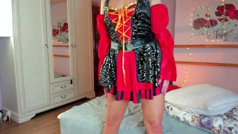 Video of a person in a red and black medieval-style costume with puffy sleeves, standing in a bedroom with white furniture and fairy lights.
