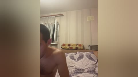 Media: Video of a man with short black hair, shirtless, sitting on a bed with a patterned white and blue duvet, in a dimly lit, simple bedroom with a window, a few clothes hanging on a rack, and a small shelf.
