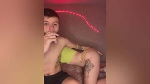 A young man with short brown hair, wearing a lime green tank top and beige underwear, lies on a bed with floral sheets. He has tattoos on his arms and legs. A red LED strip light illuminates the room.