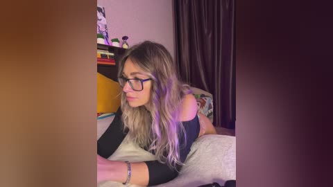 Video of a blonde woman with glasses lying on a bed, wearing a black top, in a dimly lit bedroom with purple curtains and a bookshelf.