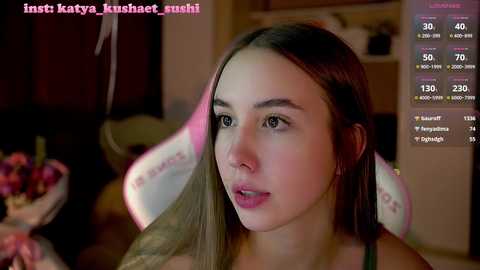 Media: A video of a young woman with long brown hair and fair skin, wearing a black top, in a dimly lit room. A live stream screen shows her stats.
