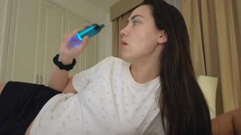 Video of a pregnant woman with long black hair, wearing a white t-shirt, holding a blue water bottle, sitting on a bed in a beige room with white curtains.