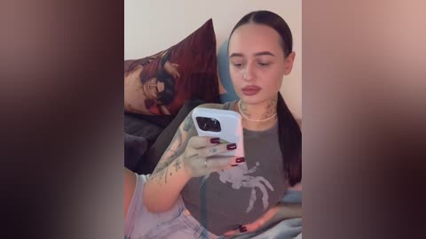 Media: A young woman with dark hair, tattoos, and a gray shirt takes a selfie in a dimly lit bedroom, surrounded by pillows and a bed.