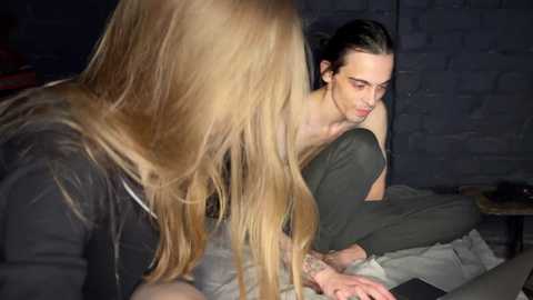 Media: Video of a blonde woman with long hair, wearing a black top, sitting on a bed, next to a man with dark hair, wearing green pants, looking intently at a smartphone.