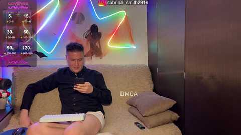 Video of a man with short hair, wearing a black shirt and shorts, sitting on a beige couch, using a tablet, surrounded by colorful neon lights and cushions.