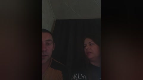 Video of a dimly lit room featuring two people, a man with short hair and a woman with long hair, both looking serious, against a dark curtain backdrop.