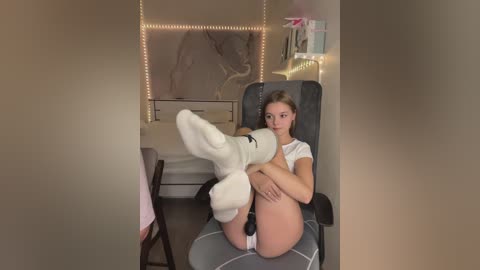 Media: Video of a young woman with fair skin and light brown hair, wearing a white t-shirt, sitting on a grey chair. She is barefoot and has a stuffed animal on her feet. The room has beige walls, a bed, and a TV with a pink bow.