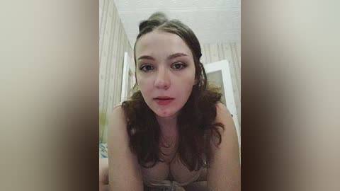 Media: Video of a young woman with fair skin, wavy brown hair, and light makeup, wearing a beige lace bralette, standing in a hallway with striped wallpaper and a white ceiling.