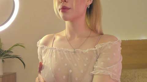 Media: Video of a fair-skinned woman with blonde hair, wearing a sheer white polka-dot top, exposing her breasts, standing indoors.