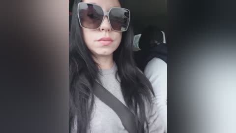 Video of a young woman with long, straight black hair, wearing large, square sunglasses and a gray sweater, seated in a car with a black seatbelt across her body.