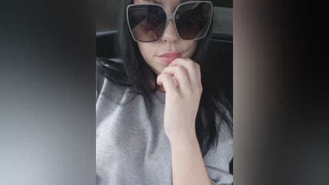 Media: A video of a young woman with long, straight black hair, wearing oversized, square, dark sunglasses and a grey hoodie, with her right hand to her chin, seated in a dark vehicle.