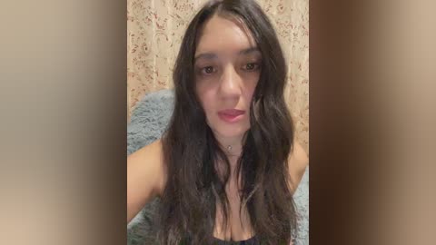 Video of a young woman with long, wavy dark hair, fair skin, and light makeup, wearing a black tank top, against a beige floral wallpaper background.