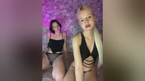 Video of two women in provocative black lingerie, sitting against a stone wall with purple lighting. One has long blonde hair, the other dark hair.