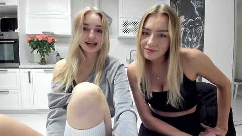 Media: Video of two blonde, slim young women in casual gray and black athletic wear, seated in a modern white kitchen with a blackboard wall and floral arrangement.