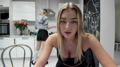 Media: Video of a blonde woman in a black bra, leaning forward in a modern kitchen, while a woman in a grey hoodie and black skirt prepares food.