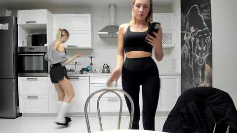 Media: Video of a modern kitchen with two women: one in a black sports bra and leggings, taking a mirror selfie, and the other in a grey top and black shorts, dancing.