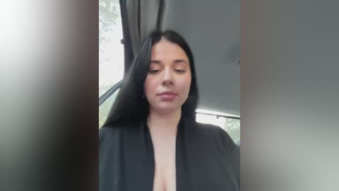 Media: A video of a young woman with long, straight black hair, wearing a black robe, standing inside a car with a window view of greenery outside.