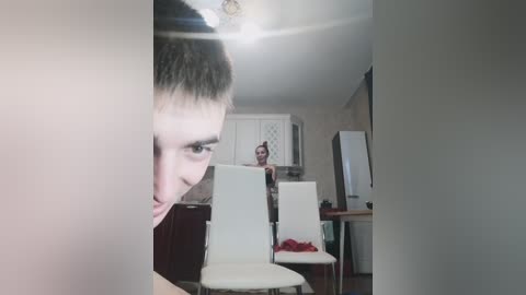 Video of a young woman with short brown hair, standing in a modern kitchen, partially obscured by a vertical object. The kitchen has white cabinets, a white refrigerator, and a red chair.