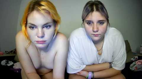 Media: Video of two young, fair-skinned women with dyed hair (one orange, one brown) posing nude, leaning forward, against a plain background.