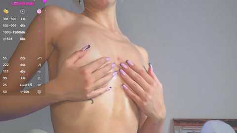 Video of a topless woman with light skin, medium-sized breasts, and manicured nails, covering her nipples with her hands. Background shows a simple room with a gray wall and a wooden table.