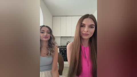 Media: Video of two young women in a modern kitchen: one with long, straight brown hair in a pink top, the other with wavy, light blonde hair in a light blue top.