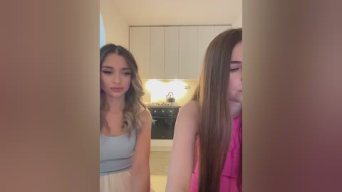 Media: Video of two young women in a modern kitchen. One wears a light blue tank top, the other a pink sleeveless top. Kitchen features white cabinets, black stove, and beige floor.