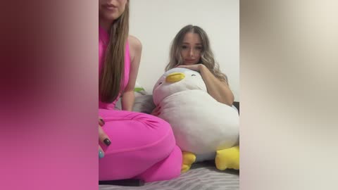 Media: Video of a young woman with long brown hair, wearing a bright pink dress, sitting on a bed with a white plush duck and a grey blanket, in a simple, lightly-lit room.