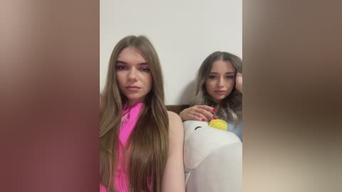 Media: Video of two young women with long, straight brown hair, one in a bright pink sleeveless top and the other in a light blue sweater. They stand against a beige wall, with the background blurred.