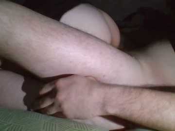 A video captures two close-up, intertwined legs of fair-skinned individuals, likely male, in a dimly lit, intimate setting. The background is blurred, focusing on the skin textures and shadows.