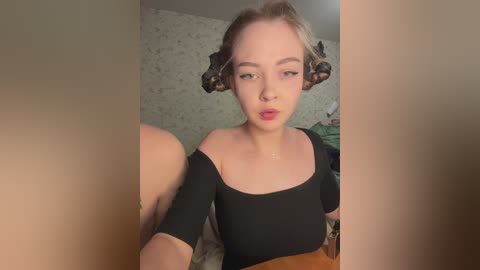 Video of a young, fair-skinned woman with blonde hair styled in buns, wearing a black off-shoulder top, in a dimly-lit room with floral wallpaper.