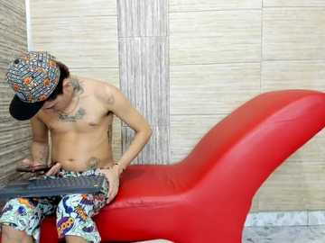 Video of a shirtless, tattooed man with short dark hair, wearing a colorful hat and patterned pants, typing on a keyboard while sitting on a red chair against beige tiled walls.