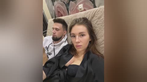 Media: Video of a man in a white hoodie and a woman in a black top, both looking tired, lying on a beige bed with floral pillows in the background.
