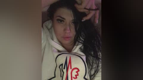 Video of a young woman with long, dark hair, light skin, and a small nose, wearing a white hoodie with red and black graphics, lying on a bed, looking serious.