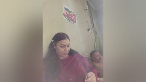A video showing a young woman with long black hair, wearing a maroon sweater, sitting on a bed with a man, both appearing to be in a domestic setting.