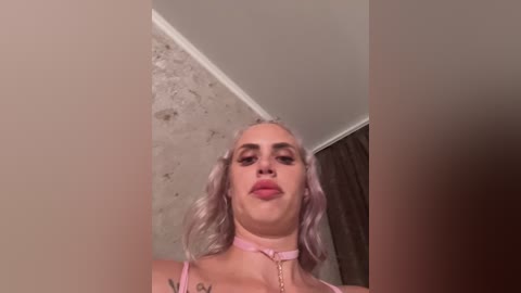 Media: Video of a woman with platinum blonde hair in a pink choker, wearing a light pink top, standing in a room with exposed concrete walls and a window partially covered by brown curtains.