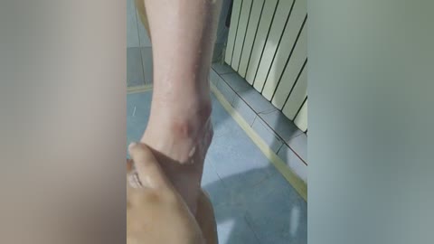 A video showing a person with fair skin and a small red mark on their lower leg being massaged by another person with darker skin. The background features a tiled wall and a blue floor.