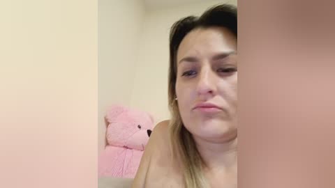 A video of a light-skinned, plus-sized woman with shoulder-length blonde hair, looking sad, standing in a corner with a pink teddy bear on a beige wall.