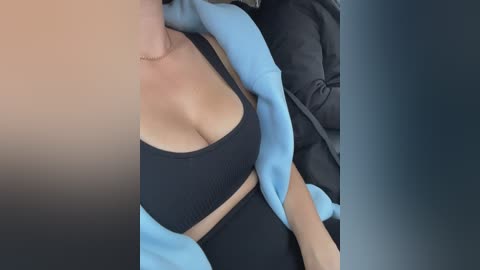 Video of a woman with light skin, wearing a black sports bra and light blue jacket, reclining in a car seat, with a blurred background of dark fabric.