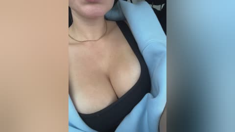 Video of a light-skinned woman with medium-sized breasts, wearing a black bra and light blue jacket. Her lips are visible, and she has a thin gold necklace on. Background is blurred, suggesting an indoor setting.