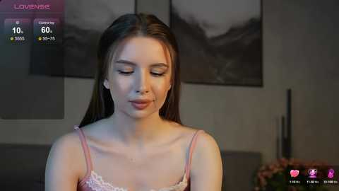 Media: Video of a young Caucasian woman with light skin, straight brown hair, wearing a pink lace camisole, in a dimly lit room with a dark abstract painting on the wall.