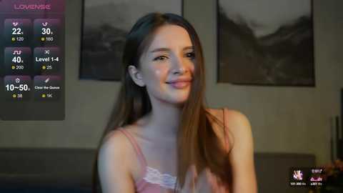 Media: Video of a young woman with long brown hair, smiling, wearing a pink lace bra, in a dimly lit room with blurred mountain scenery on the wall.