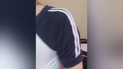A video of a person's upper torso, wearing a navy blue and white short-sleeved athletic shirt with stripes on the sleeves, standing against a blurred beige wall.