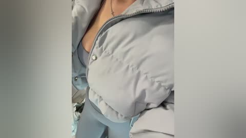 Media: Video of a woman's torso in a light blue puffy jacket, revealing a cleavage, with a white necklace, and a blurred background.