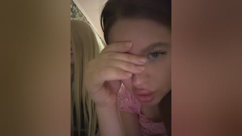 A close-up video of a young Caucasian woman with fair skin, blue eyes, and long blonde hair. She wears a pink lace top and touches her forehead with her right hand, looking contemplative. The background includes a mirror and a floral patterned wall.
